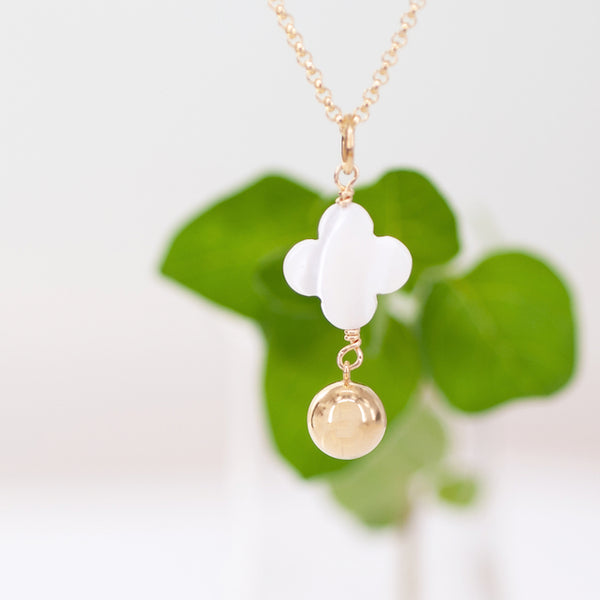 Necklace with a white four-leaf clover pendant and gold ball on a light background with green leaves.
